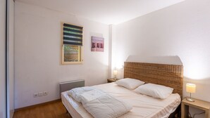 A bedroom with a bed, a nightstand, a lamp, a window with blinds, and a radiator. - Apartment Near Plage Moutchic Lake (Lacanau)