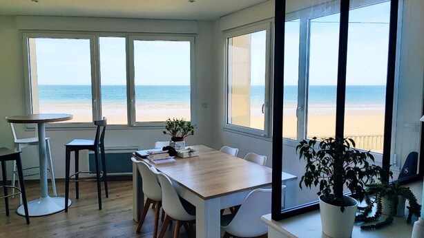 Ein Essbereich mit Holztisch und -stühlen, großen Fenstern mit Blick auf den Strand und Topfpflanzen.