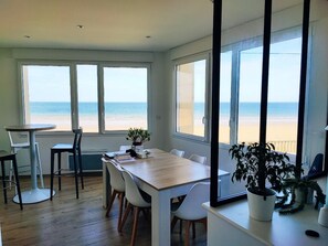 A dining area with a wooden table and chairs, large windows with a beach view, and potted plants. - Apartment by the Sea (Grandcamp-Maisy)