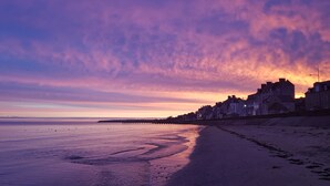 Ein Strand mit einer Reihe von Häusern entlang der Küste, einer Mole, die sich ins Wasser erstreckt, und einem Sonnenuntergang im Hintergrund.