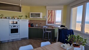 Eine moderne Küche mit Einbauofen, Mikrowelle und Kaffeemaschine. Es gibt einen Essbereich mit Tisch und Stühlen sowie ein großes Fenster mit Blick auf den Strand.