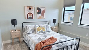 Un dormitorio con cama, dos mesitas de noche, una ventana y dos cuadros.