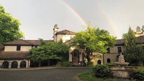 Un cortile con un edificio caratterizzato da un tetto in tegole e finestre ad arco, circondato da vegetazione rigogliosa e una fontana in pietra.