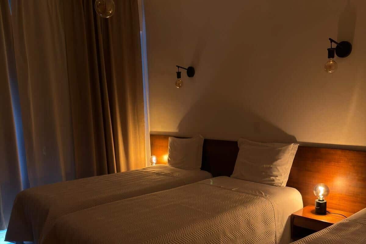 A hotel room with two beds, a wooden headboard, a nightstand with a lamp, and a window with curtains.