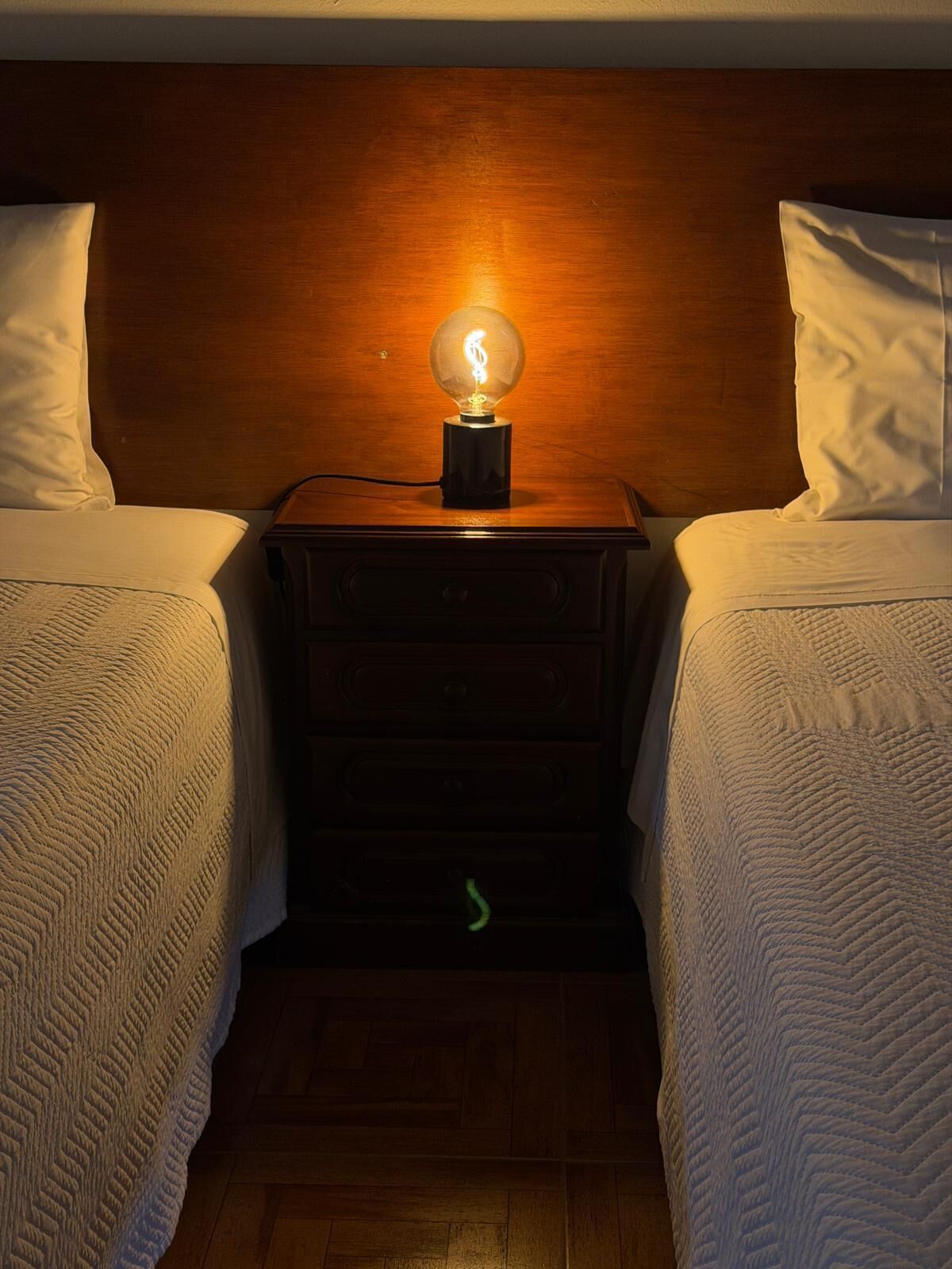Habitación de hotel con dos camas, un cabecero de madera, una mesita de noche con lámpara y una luz verde en el suelo.