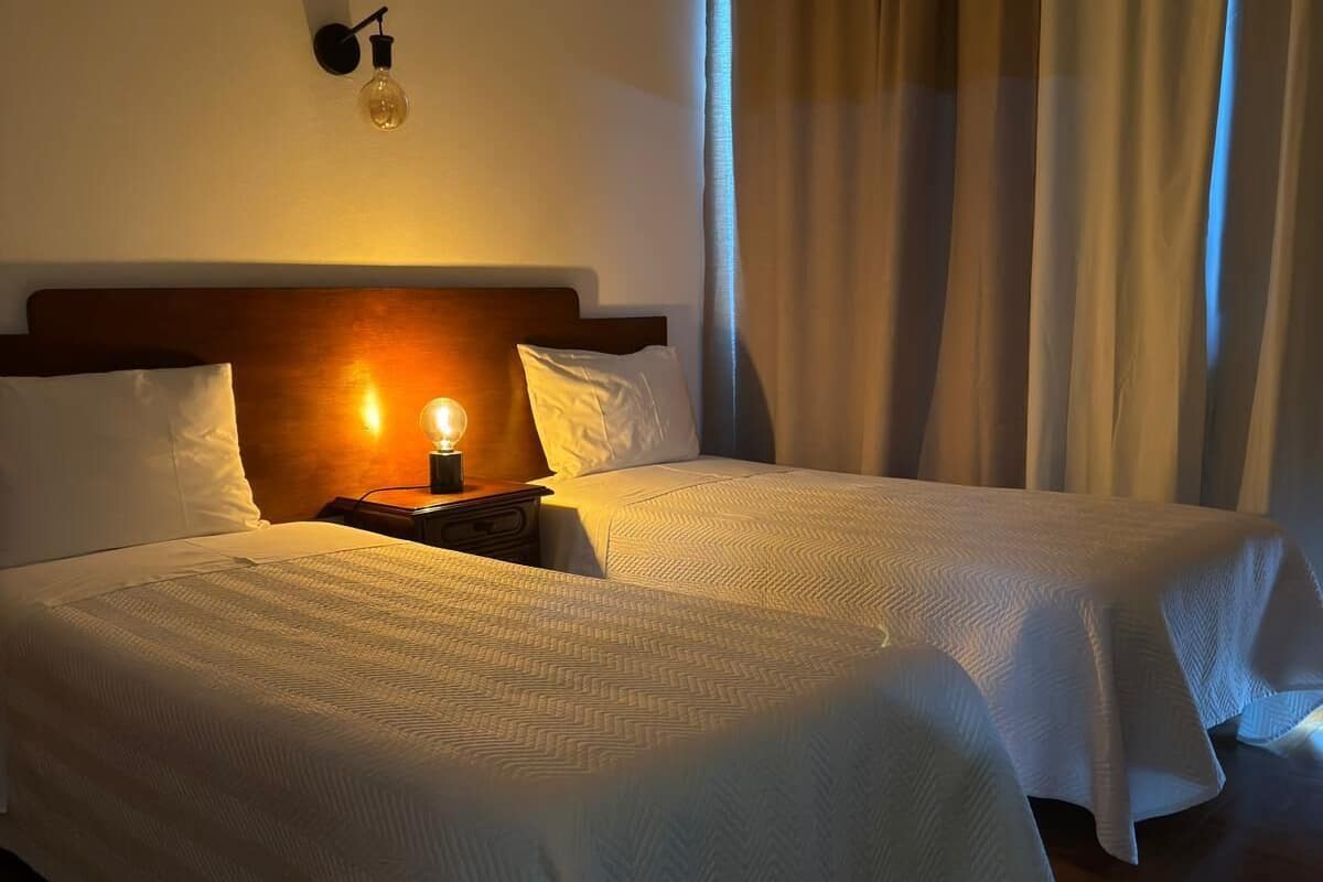 A hotel room with two beds, a nightstand with a lamp, and a window with curtains.