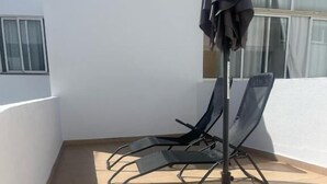 Un piccolo spazio esterno con pavimento piastrellato, due poltrone nere, un ombrellone e una finestra con tende.
