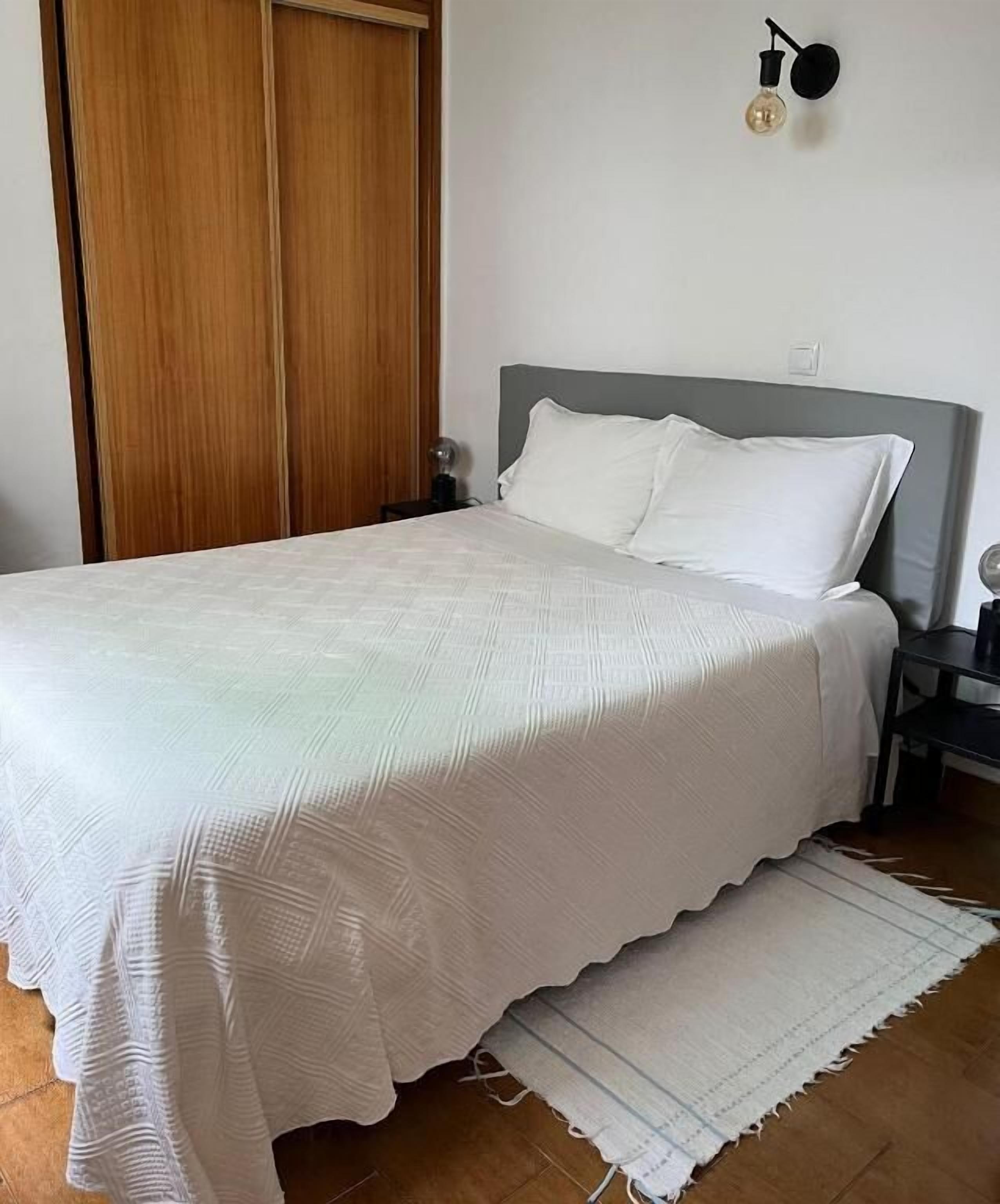 A neatly made bed with a white bedspread and pillows, a wooden wardrobe, and a wall-mounted light fixture.