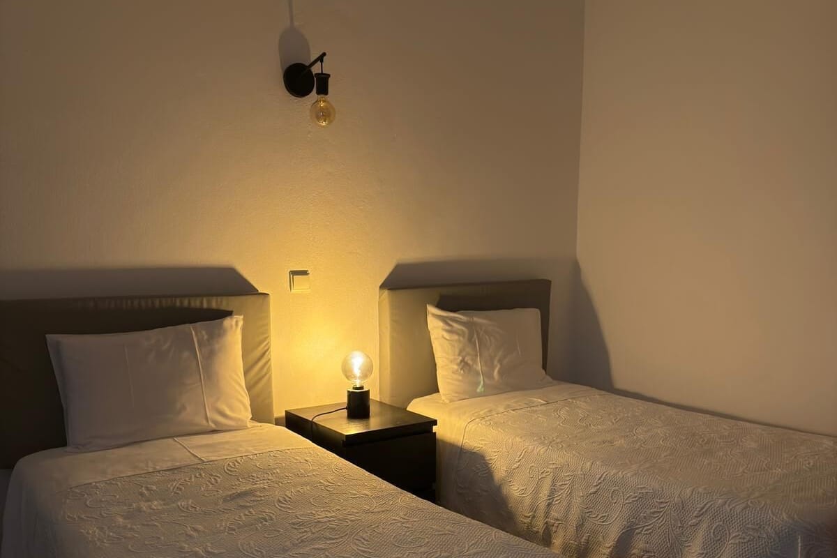 A room with two beds, each with a headboard and a bedside lamp.