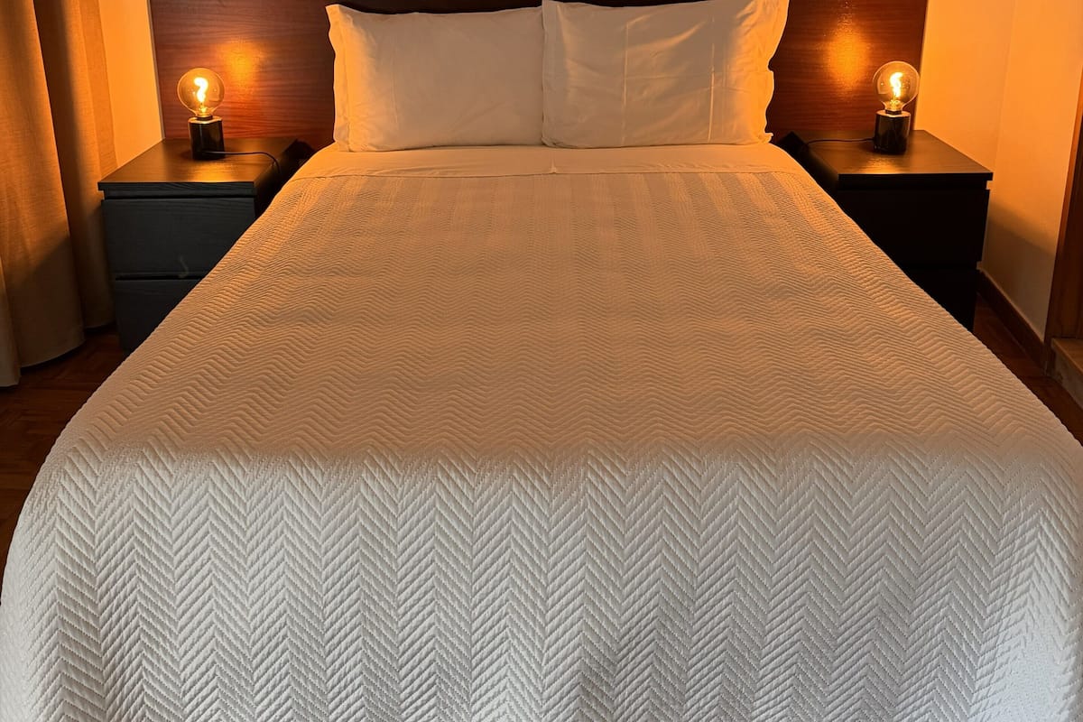 A neatly made bed with a white quilt, two bedside lamps, and a wooden headboard.