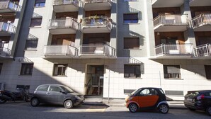 Um edifício residencial com vários andares, varandas, um carro estacionado na frente e uma motocicleta à esquerda.