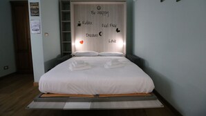 Uma cama com colchão e almofadas brancas, cabeceira de madeira com palavras motivacionais e uma pequena prateleira acima da cama.