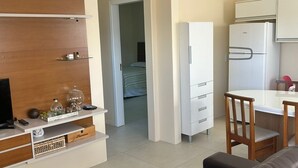 Un espacio habitable compacto con sofá, televisor, zona de cocina que incluye un refrigerador y una mesa de comedor, además de un dormitorio pequeño visible a través de una puerta abierta.