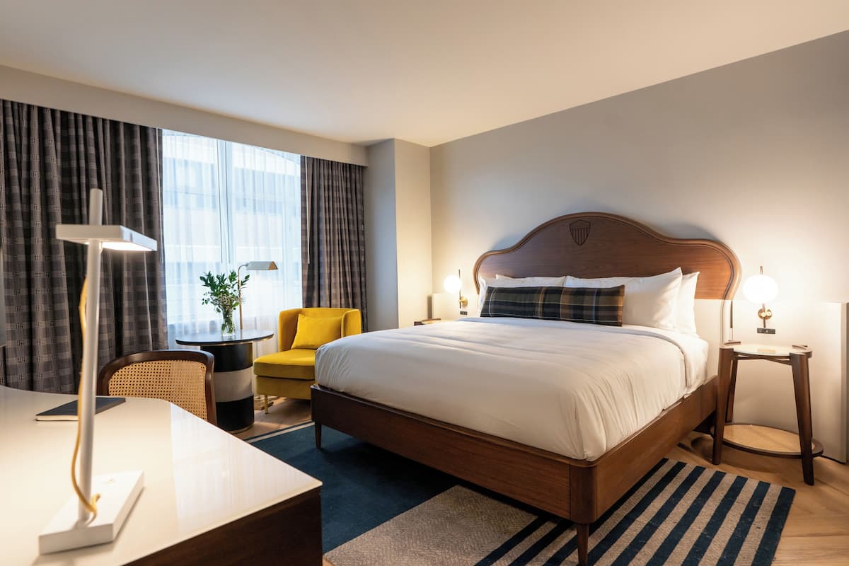 A hotel room with a large bed, a desk, a chair, a lamp, and a view of the city.