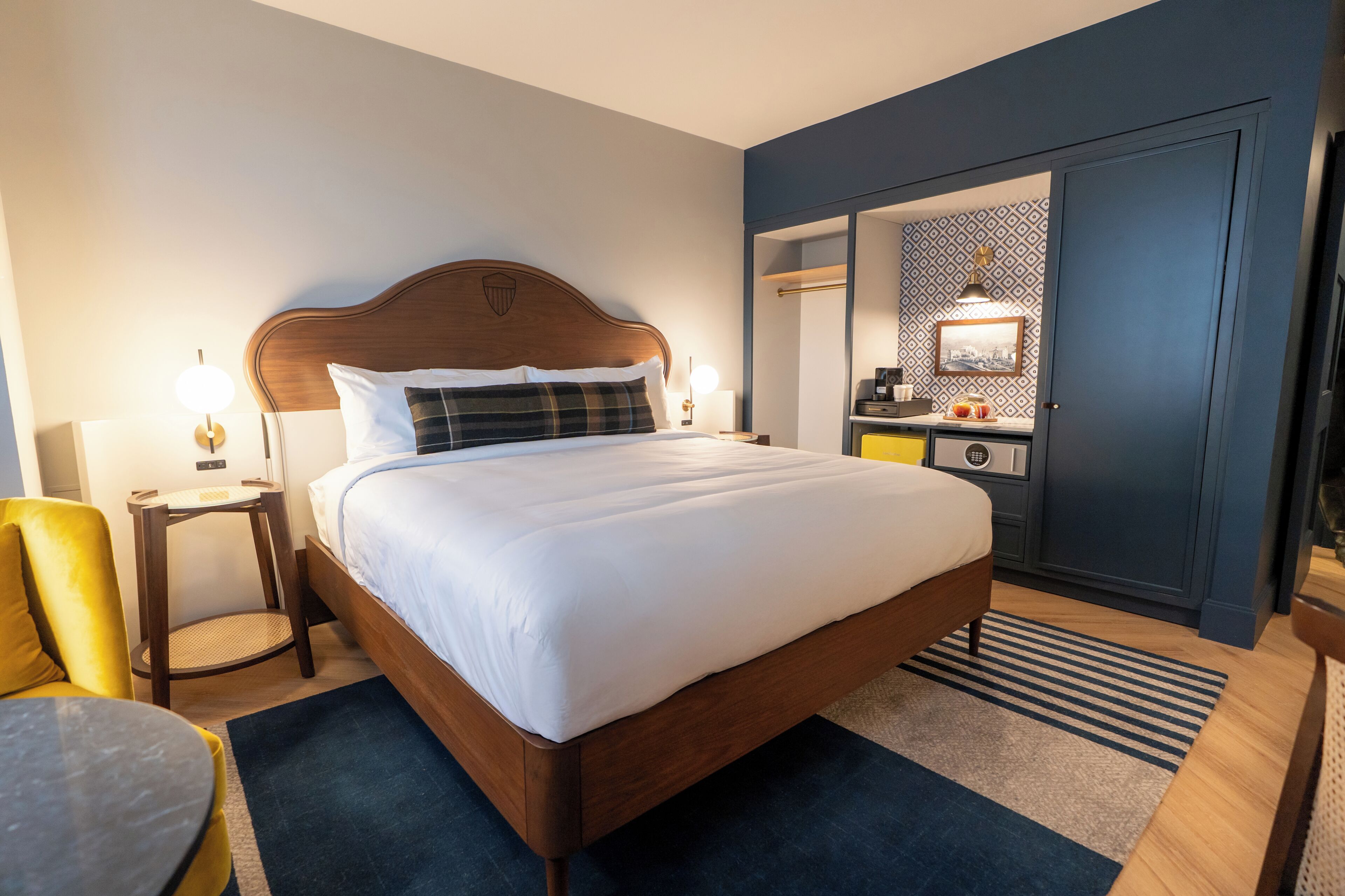 A hotel room with a bed, bedside table, lamp, chair, and a small table with a lamp.