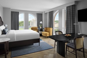 A hotel room with a large bed, a desk, a chair, a TV, and a view of the city.