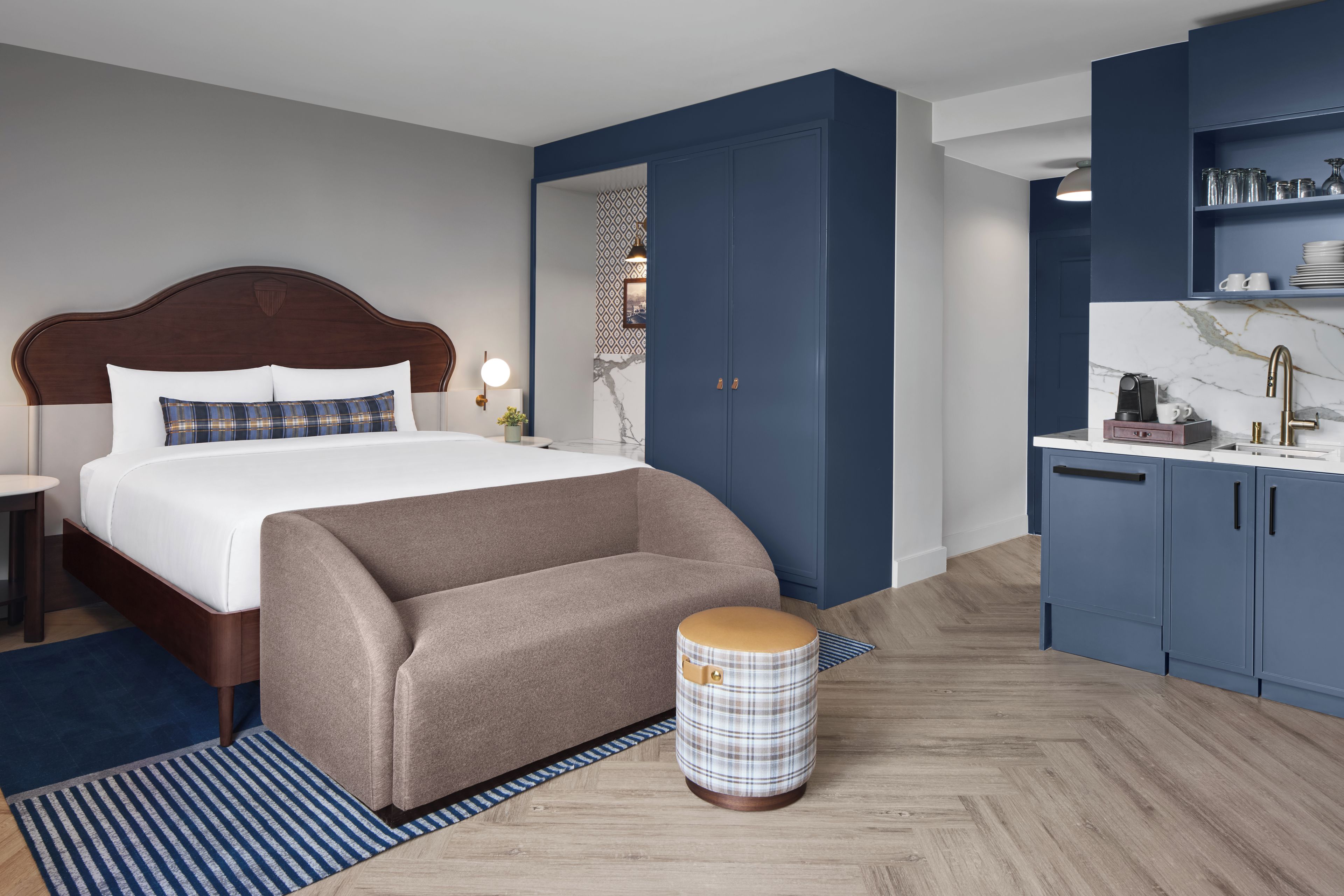 A modern hotel room with a large bed, a small sofa, a round ottoman, a desk with a chair, and a built-in kitchen area.