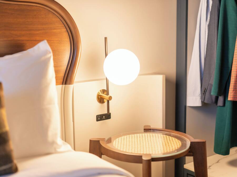 Habitación de hotel con cama, mesita de noche con lámpara, una silla y espejo adosado a la pared.