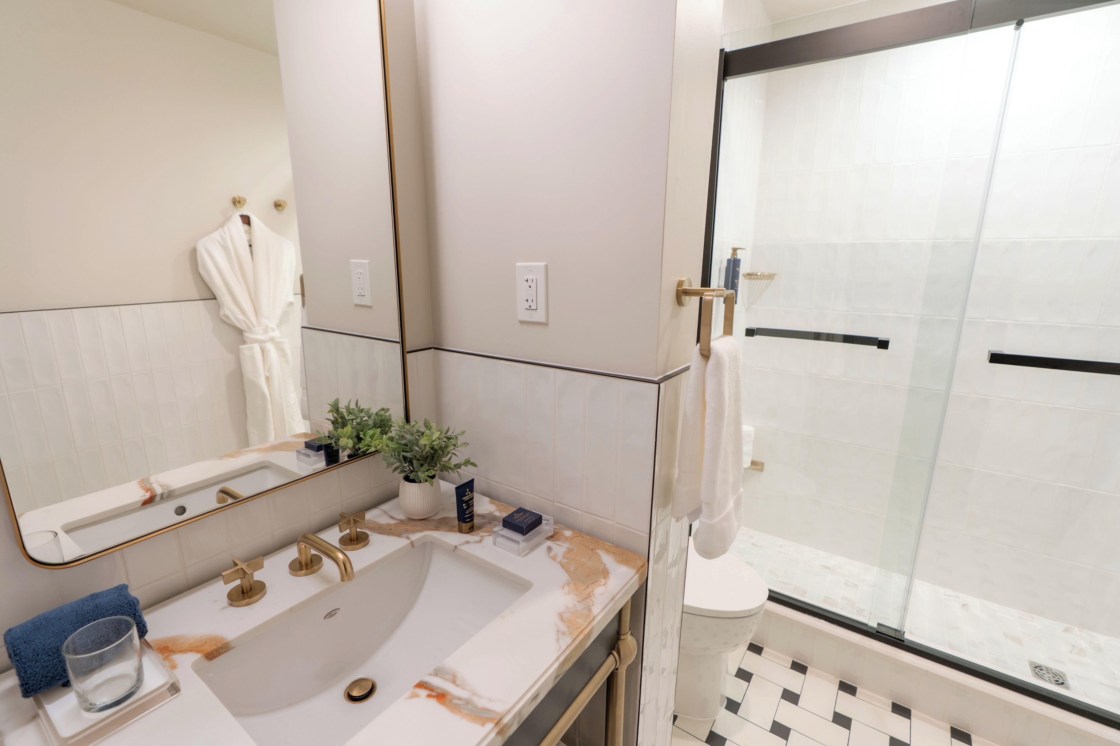 A bathroom with a large mirror, a sink with gold fixtures, a potted plant, and a glass shower enclosure.