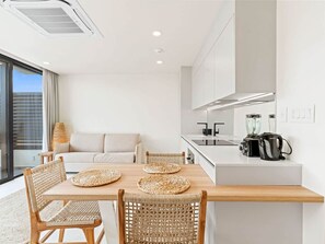 A modern kitchen with a central island, a dining area with woven chairs, and a sofa in a bright room with large windows.