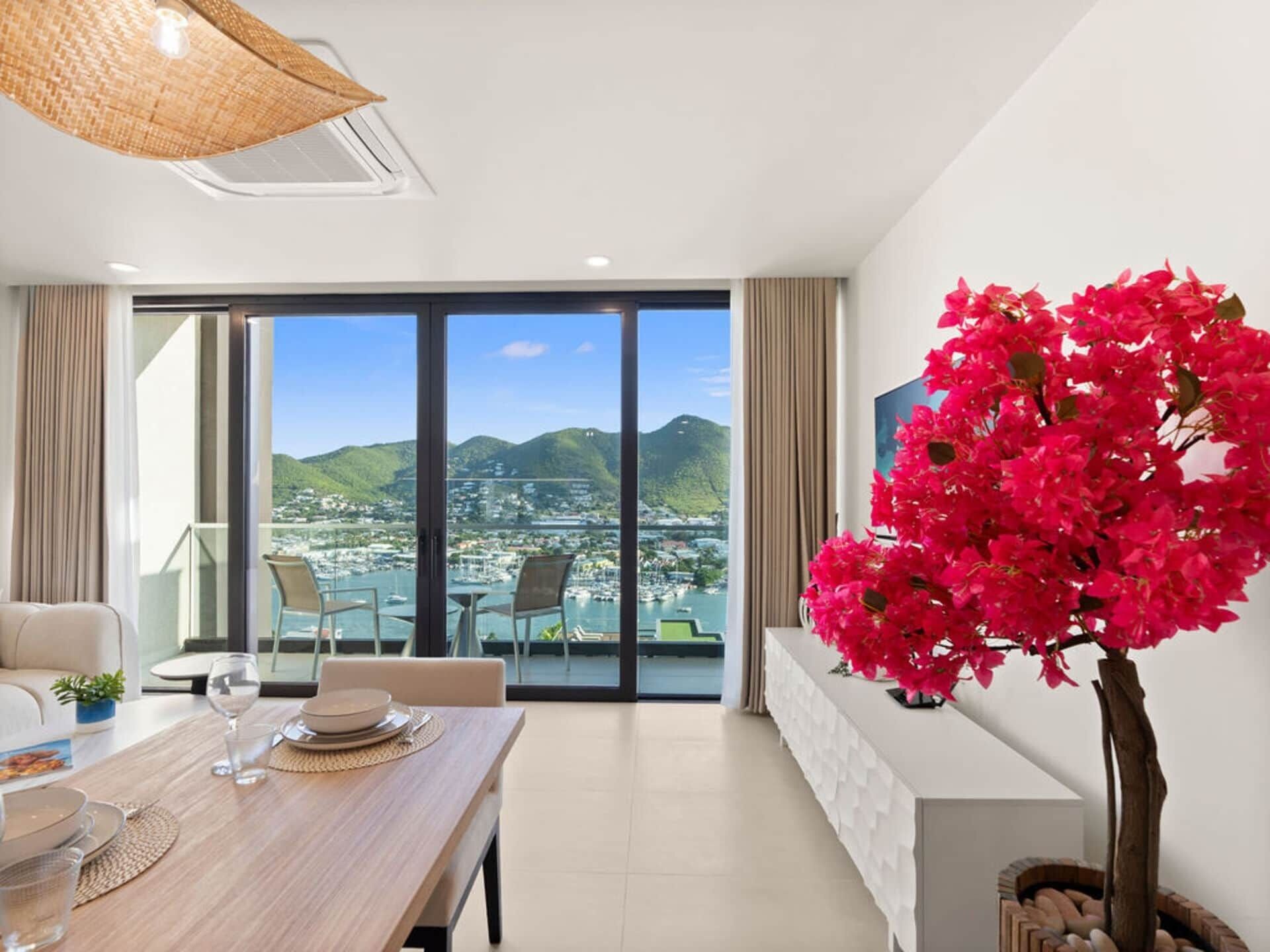 A modern hotel room with a dining area, a large window with a city and mountain view, and a potted pink bougainvillea plant.