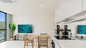 A modern kitchen with a dining area, featuring a large island, white cabinetry, and two wall-mounted TVs displaying a coastal scene. - D417 - One Bedroom Apartment with Lagoon View (Cole Bay)