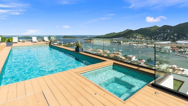 Una piscina infinita en la azotea, con terraza de madera y barandillas de vidrio, con vistas a un puerto deportivo y al paisaje urbano.