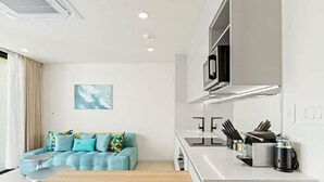 Una cocina moderna con encimera blanca, estufa integrada y microondas. En la zona de estar contigua, hay un sofá con cojines azules.