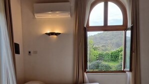 A bedroom with a wooden floor, a bed with a quilted cover, a window with curtains, and a wall-mounted air conditioner. - Relax nel Verde 2 - One Bedroom Apartment, Sleeps 3 (Calafuria)