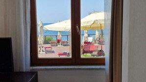 A room with a large window overlooking a beach area with umbrellas and lounge chairs. - Risveglio vista mare 2 (Calafuria)
