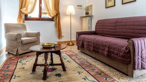 Een gezellige woonkamer met een gestreept fauteuil, een ronde houten tafel met een schaal, een paarse bank, een kamerkleed met dessin en een vloerlamp.