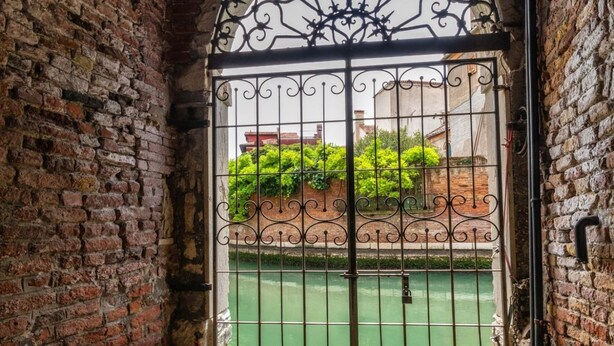 Een bakstenen muur met een metalen poort die uitkomt op een binnentuin met een groene ondergrond.