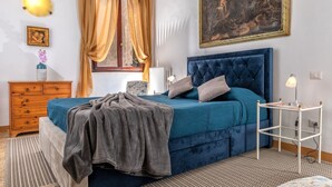 Een slaapkamer met een groot bed, een nachtkastje, een kaptafel, een raam met gordijnen en een schilderij aan de muur.