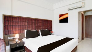 A hotel room with a large bed, a nightstand with a lamp, a wall-mounted air conditioner, and a painting of a sunset.