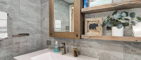 Une salle de bain avec un lavabo blanc, un miroir encadré en bois et une étagère contenant des articles de toilette et un tableau encadré.