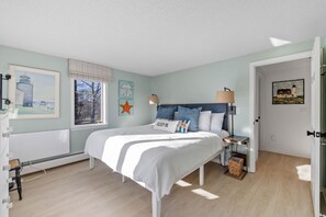 Une chambre à coucher avec un lit, des tables de chevet, une fenêtre avec des stores et des œuvres d’art mural.
