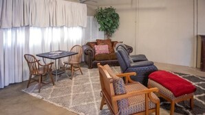 Ein Wohnzimmer mit einem braunen Sofa, einem blauen Sessel, einem Holztisch und zwei Holzstühlen. In der Ecke steht eine große Pflanze, und ein gemustertes Teppich bedeckt den Boden.