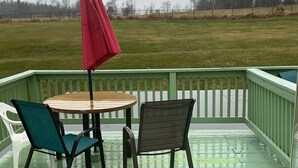 Een groen terras met een ronde tafel en twee stoelen, een rode parasol en uitzicht op een grasveld.