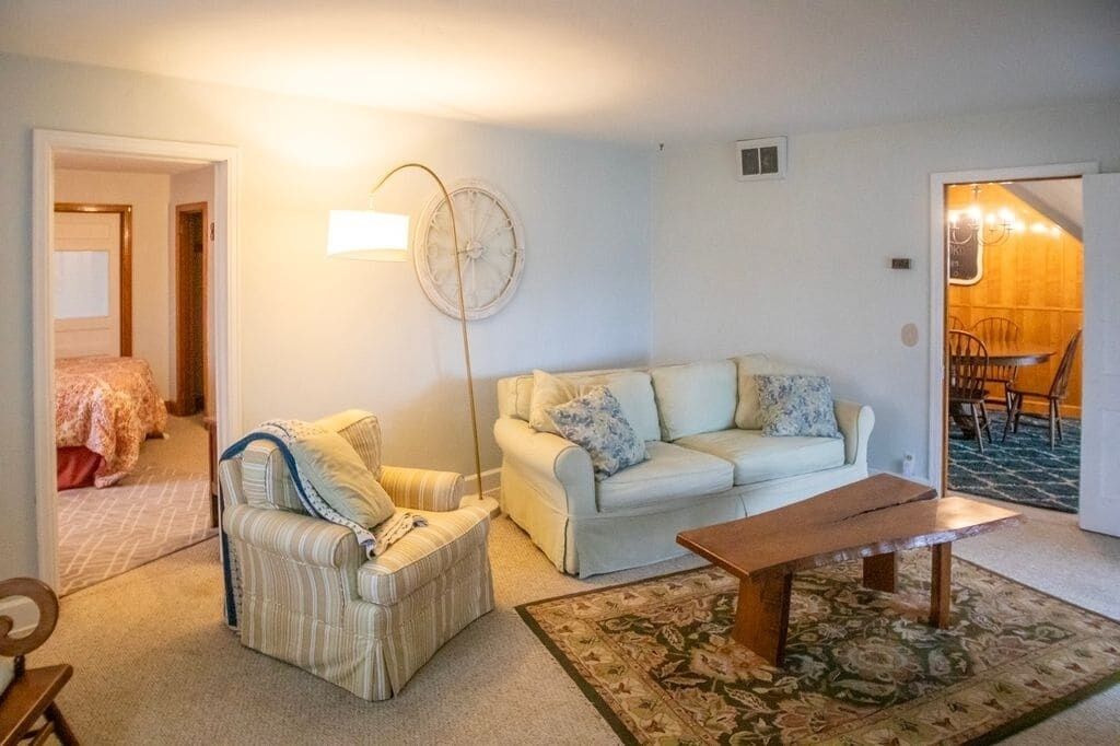 A living room with a sofa, armchair, and a coffee table. There is a lamp and a clock on the wall.