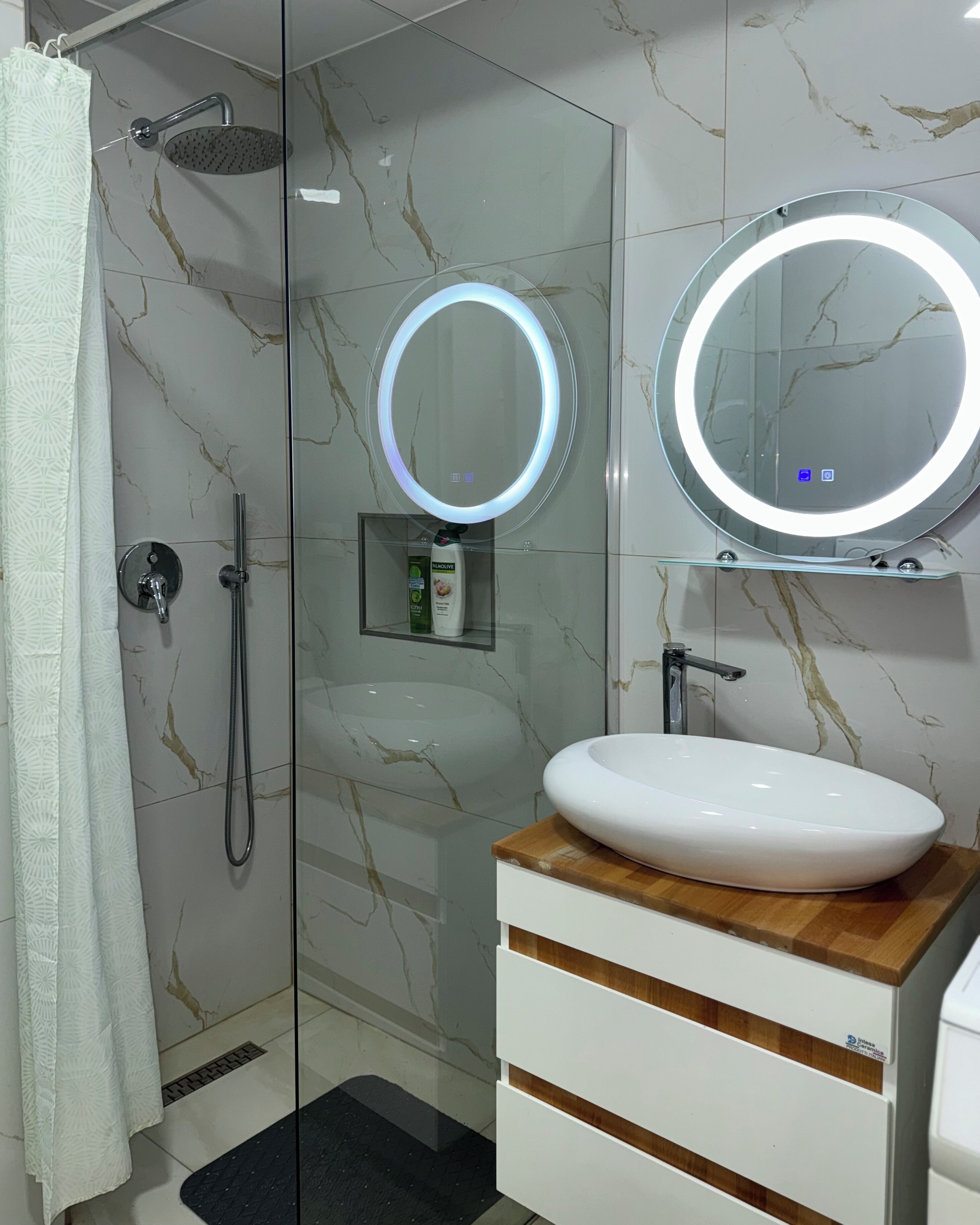 Ein modernes Badezimmer mit einer in Glas abgeschlossenen Dusche, einem weißen Waschbecken mit Holzplatte und zwei an der Wand befestigten Spiegeln mit beleuchteten Rahmen.