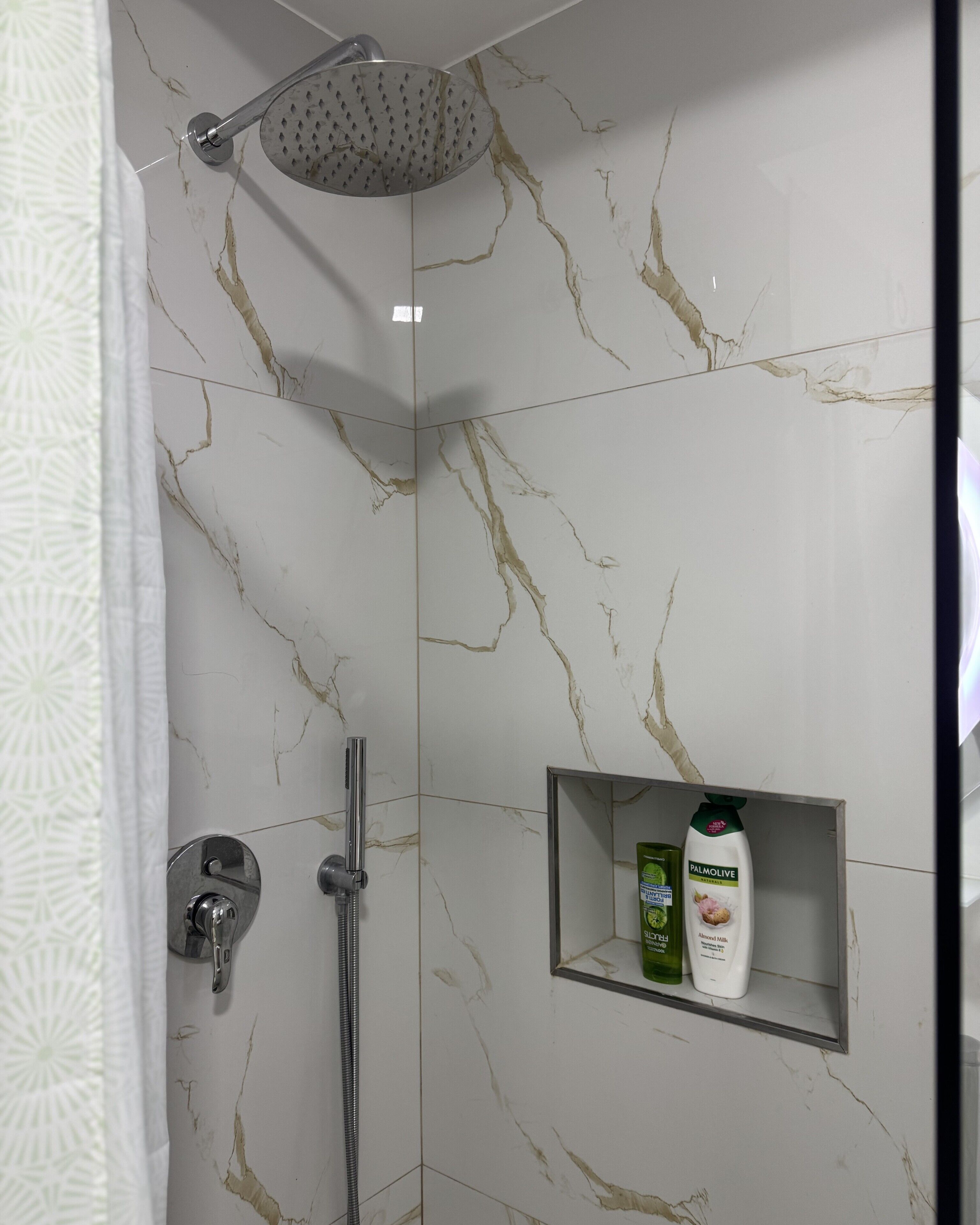 A shower with a handheld showerhead and a handheld shower attachment. There is a shelf with two bottles of Palmolive shampoo and conditioner.