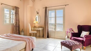 Une chambre à coucher avec un lit, un bureau avec une chaise, une coiffeuse avec un miroir, une fenêtre avec des rideaux, un fauteuil violet et une petite table avec des fleurs.