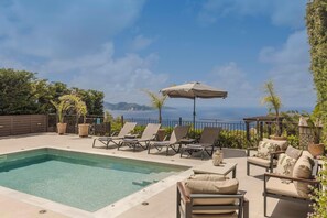 A pool area with lounge chairs and an umbrella, surrounded by greenery and overlooking a body of water. - Vasilikades - Three Bedroom Villa, Sleeps 6 (Fiskardo)
