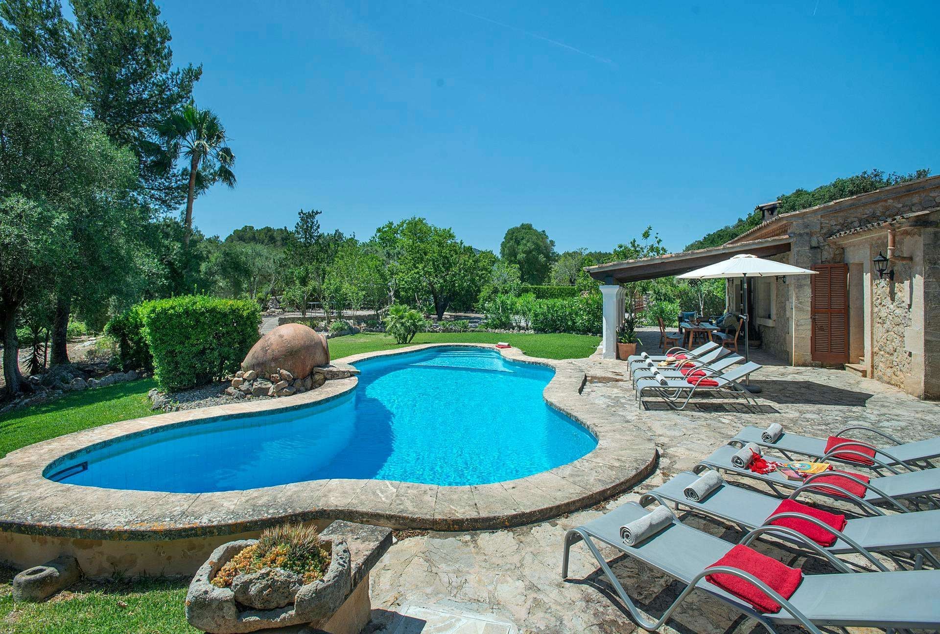 Piscina com borda curva, cercada por um deck de pedra. Há cadeiras de descanso com almofadas vermelhas e uma pérgula com guarda-sol.