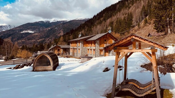 Una xalet de fusta amb una cadira bressol de fusta i un botlle de fusta en un paisatge nevà amb muntanyes al fons.
