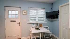 Una habitación pequeña y bien iluminada, con una puerta blanca, una ventana con persianas, un televisor montado, una mesa blanca con una silla y una planta pequeña.
