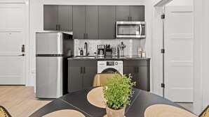 Una cocina moderna con armarios grises, un refrigerador de acero inoxidable, un microondas y una cafetera. Hay una mesa de comedor con una planta en maceta y una silla.