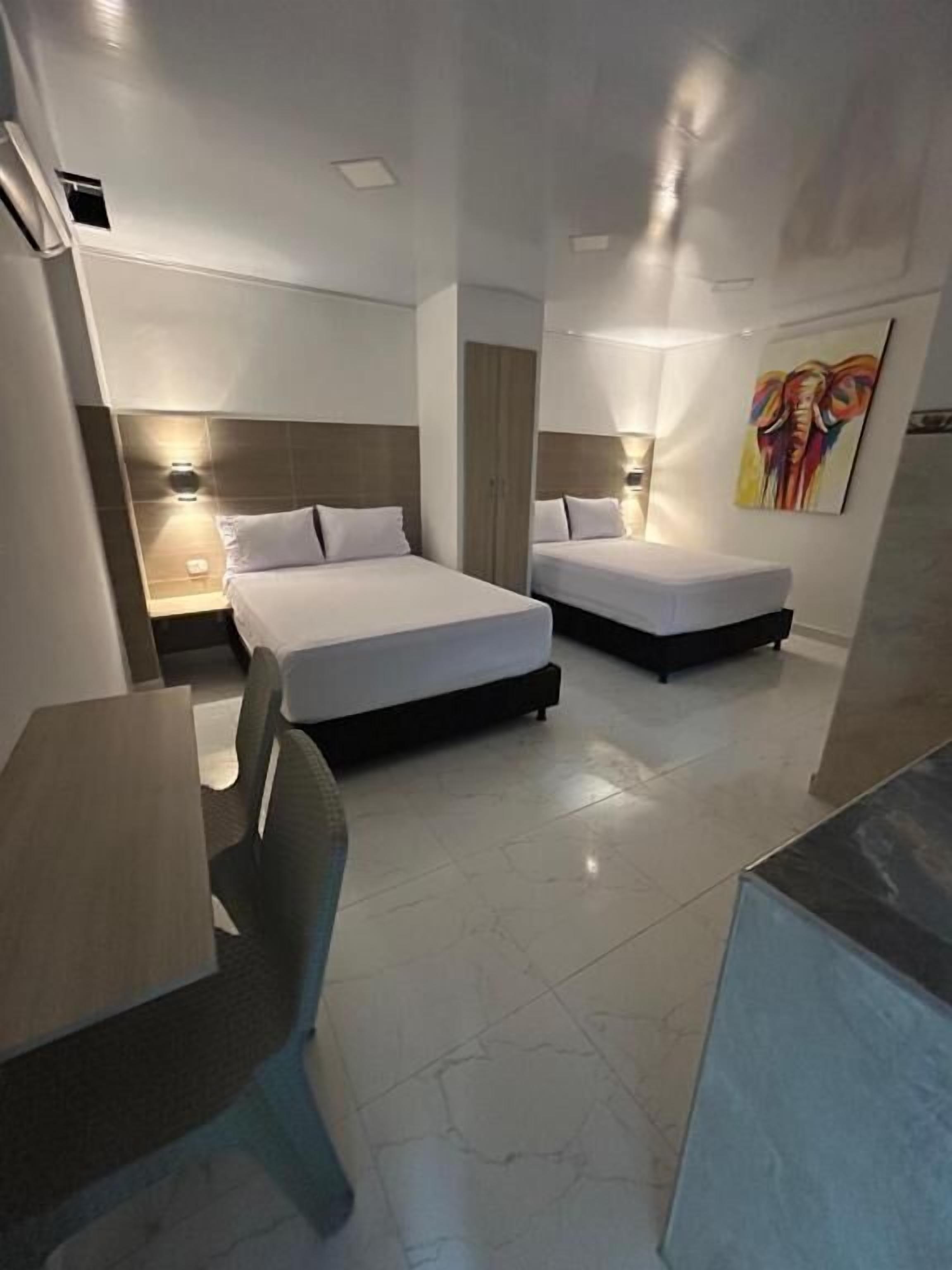 Habitación de hotel con dos camas, un escritorio y una silla. Tiene luces de pared y un cuadro colgado en la pared.
