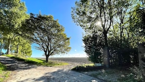 Uma trilha pavimentada que conduz a um corpo d'água, ladeada por árvores e uma construção de tijolos.
