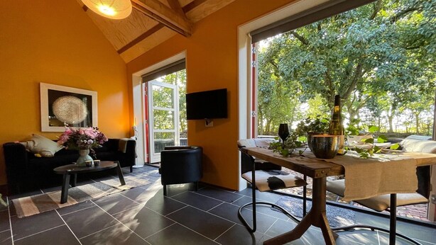 Sala de estar moderna com sofá, mesa de centro e mesa de jantar, janelas amplas com vista para árvores e uma televisão de tela plana na parede.